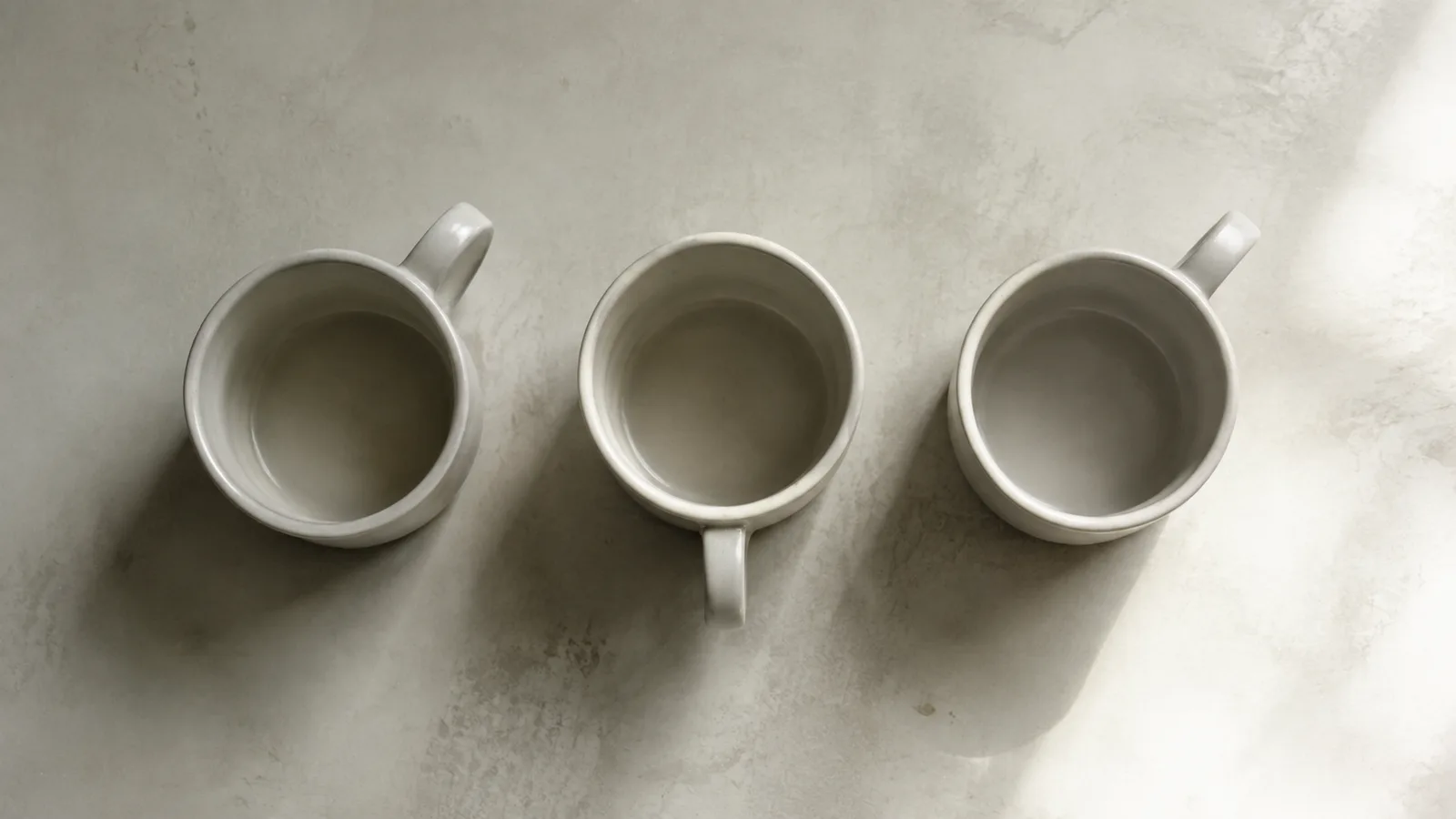 Three near-identical ceramic mugs seen from above on a pale surface, one turned away from the others, representing a buyer comparing options that look the same.